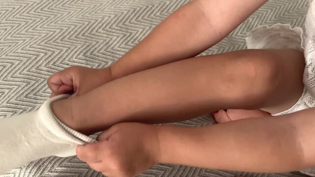 Child pulling a white sock onto one foot while sitting indoors, dressing practice, self care, daily routine, childhood learning.