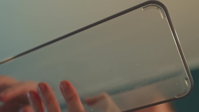 Close up of hands interacting with transparent rectangular objects reflecting light. Hands move gently over glass or plastic surfaces creating ripples. Teal wall background visible