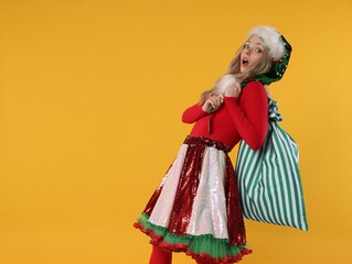 Fototapeta premium Woman dressed as elf with heavy sack on yellow background, space for text. Christmas celebration