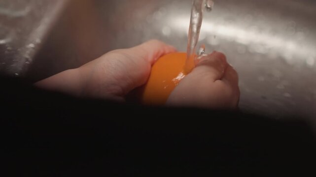 Close up of hands washing orange under running water in sink. Fruit is rotated and rinsed thoroughly with visible wet skin. Metallic kitchen setting provides context