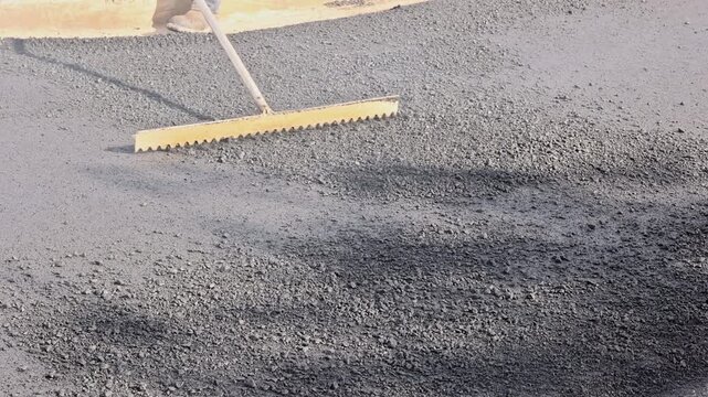 Worker uses rake to smooth fresh asphalt on road surface in city
