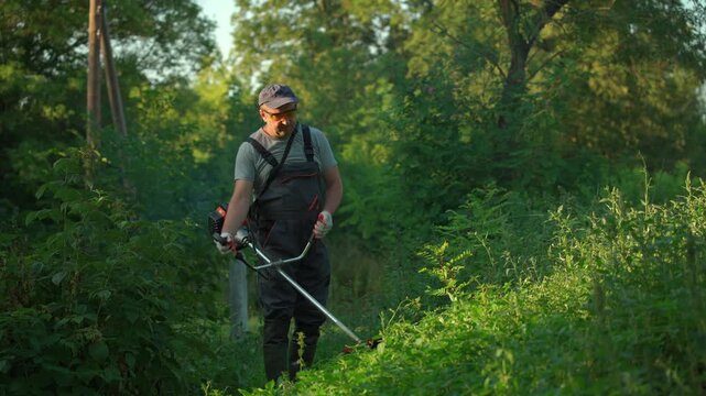 Gardener in gray overalls uses a string trimmer to clear tall grass and weeds in a lush green garden area, surrounded by trees and natural vegetation under bright sunlight