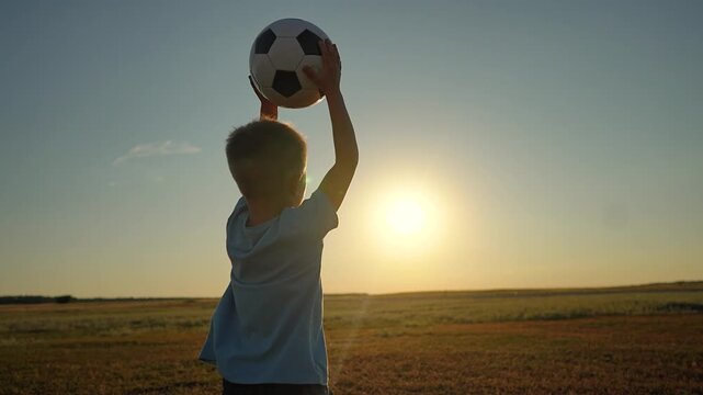 Little boy holding soccer ball stands in sunset field. Child lifts football against bright sun outdoors. Kid with ball dreams of big game. Active boy plays sport in park. Young soccer player training.