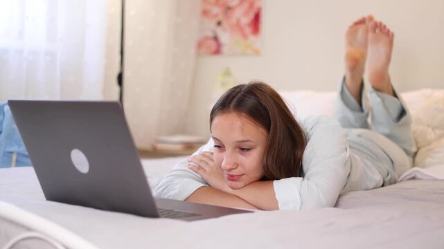 Teenage girl using laptop while lying on bed at home. Modern lifestyle, online communication and remote learning. 