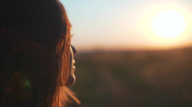 Young woman face looks at sunset in nature field. Close up of female profile with hair blowing in wind. Happy girl enjoys bright golden sun outdoors. Woman watches warm summer sunset sunlight.