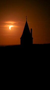 Vertical drone footage of Bouzov Castle in silhouette with a partial solar eclipse setting behind the tower in warm evening light, Czech Republic. Colors: orange, black, yellow.