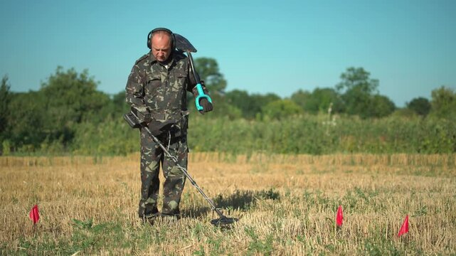Male treasure hunter in camouflage outfit uses metal detector while holding a shovel in a field marked with red flags, surrounded by greenery and clear blue sky in the background