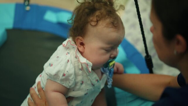 Infant child wearing a patterned romper and pacifier, lying on a blue mat, with a parent gently holding
