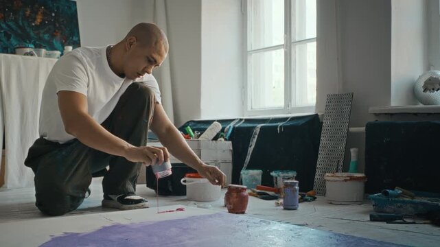 Wide shot of young adult man crouching in art studio, dripping red paint onto canvas on floor. Clip is perfect for creative industry promos and art school advertising
