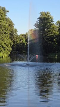 Tall water fountain spraying high in park lake creating colorful rainbow in mist surrounded by green trees on sunny summer day vertical view.