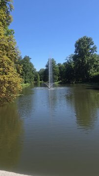 Tall water fountain spraying high in park lake creating colorful rainbow in mist surrounded by green trees on sunny summer day vertical view.