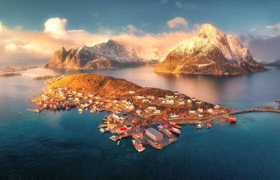 Aerial view of Reine village surrounded by fjords and snowy mountains in Lofoten, Norway. Bright winter sunlight over fishing cabins, blue sea, and dramatic coastal landscape. Top drone view