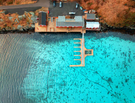 Aerial view of wooden pier, beach, transparent azure water, houses on the sea coast in summer. Sakrisoy, Lofoten islands, Norway. Top down view from drone of sea, jetty, rorbuer, orange grass. Nature