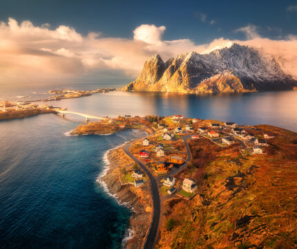 Aerial view of Reine village surrounded by fjords and snowy mountains in Lofoten, Norway. Bright winter sunlight over fishing cabins, blue sea, and dramatic coastal landscape. Top drone view