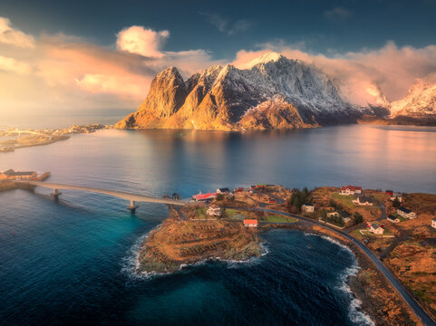 Aerial view of Reine village surrounded by fjords and snowy mountains in Lofoten, Norway. Bright winter sunlight over fishing cabins, blue sea, and dramatic coastal landscape. Top drone view