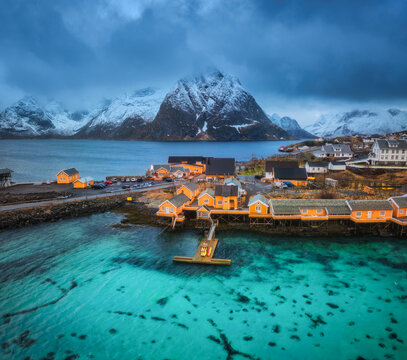 Aerial view of Hamnoy fishing village with yellow rorbuer cabins on turquoise water in Lofoten, Norway. Scenic coastal landscape with snowy mountains, wooden houses, and dramatic arctic sky. Top view