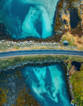 Asphalt road highway over turquoise ocean water in Lofoten, Norway. Aerial top down drone view of coastal road, clear sea bottom, rocky shore, moss and scenic arctic island landscape in autumn