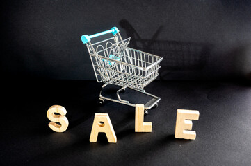 Wooden letters spelling SALE with a small metal shopping cart on a dark moody background a creative retail concept of discounts special offers and seasonal shopping. © Oksana