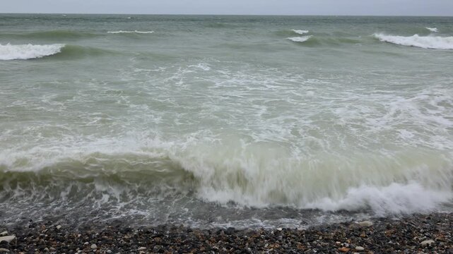 Waves rolling onto a shingle beach at Cap Blanc-Nez, France. Panning shot of green sea water, breaking surf and foam running up the pebbles on the Opal Coast on a windy grey day