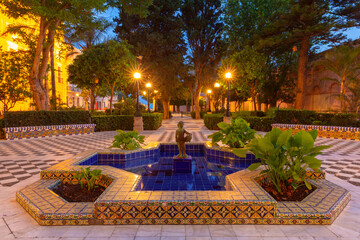 Decorative ceramic fountain surrounded by tiled benches, trees and illuminated pathways in a historic garden in Cadiz, Spain. © Kavalenkava