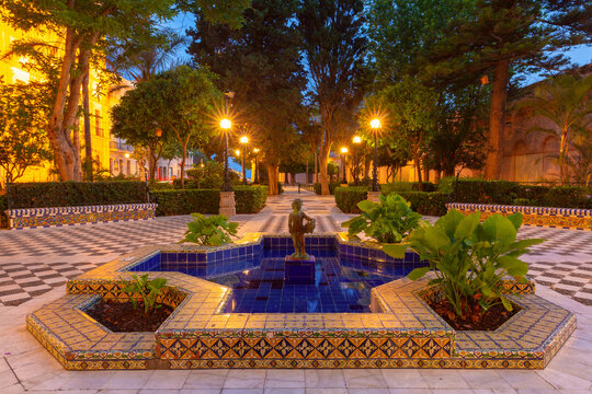 Decorative ceramic fountain surrounded by tiled benches, trees and illuminated pathways in a historic garden in Cadiz, Spain.