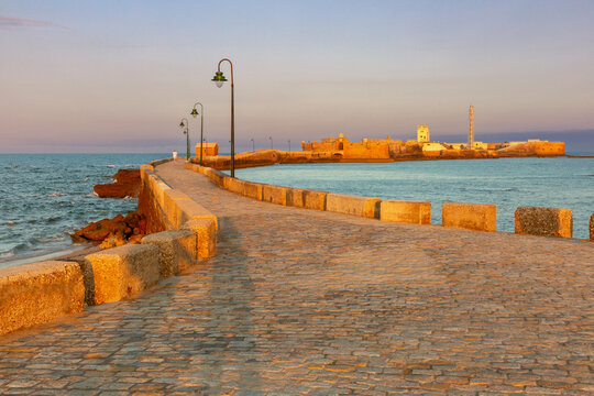 Stone causeway leading toward San Sebastian Fort across the Atlantic coast in Cadiz, Spain, illuminated by warm sunrise light.