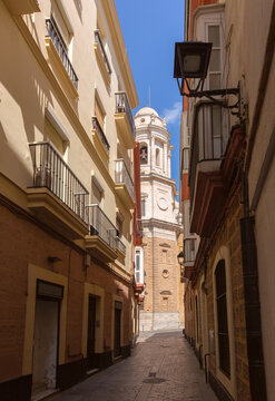 Traditional buildings with balconies frame a narrow pedestrian lane overlooking the cathedral bell tower in Cadiz, Spain.