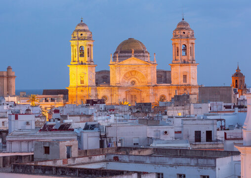 Illuminated Cadiz Cathedral rising above the white rooftops of the old town during sunset in Cadiz, Andalusia, Spain.