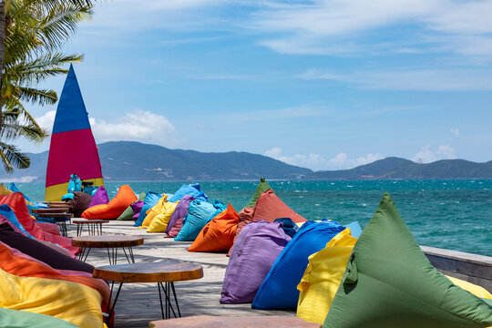 Marina beach in Nha Trang. Colorful bean bags arranged on a wooden deck overlooking a tropical beach with clear blue water and distant mountains under a bright sky. Beach club and vacation concept