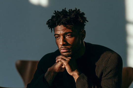 A close-up portrait of a thoughtful young Black man with textured hair, posing with hands clasped under his chin, wearing a dark sweater in a studio setting with soft natural window light and shadow.
