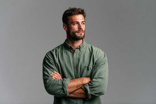 Professional studio headshot of a handsome young man with a beard wearing a green button-down shirt, standing with arms crossed looking away, isolated on a neutral gray background for corporate use.