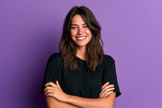 A friendly and confident young woman with wavy brown hair wearing a simple black t-shirt standing with arms crossed in a studio headshot portrait against a vibrant solid purple background for