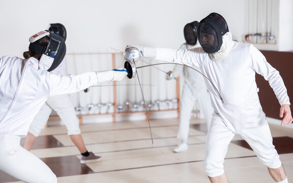 Male and female fencers practice in pairs in sports hall, wearing white gear and protective masks