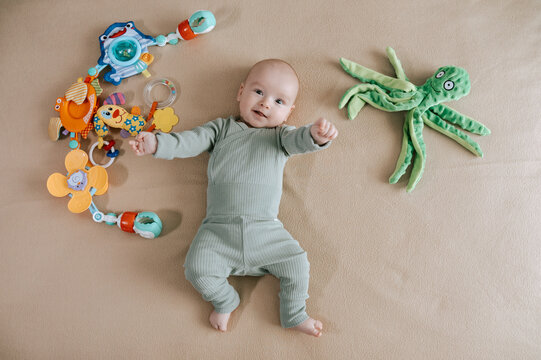 Cute 3 month baby boy lying among various playful colorful toys on beige blanket. Infant surrounded by vivid objects participating actively in sensory exploration and learning. 