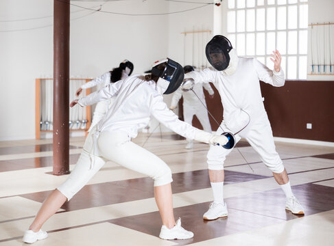 Athletes in protective equipment and helmets conduct a fencing training match in the gym. People at a group fencing lesson practice with rapier
