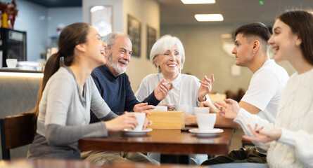 Fototapeta premium Elderly couple two young women and young man talking and drinking coffee at table in cafe