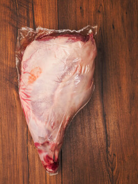 Raw lamb leg in plastic wrap rests on wooden surface. Meat shows pink flesh and red fat marbling. Scene suggests butchery, cooking, or grocery retail. Perfect for culinary, food