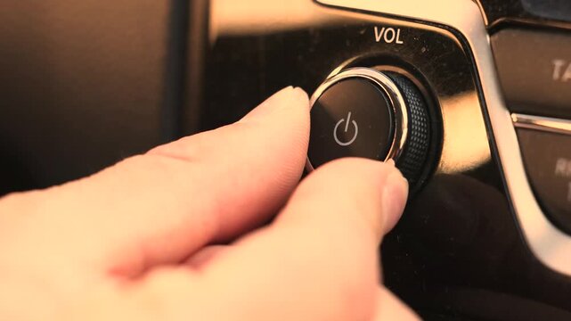 Finger turning the volume knob clockwise on a modern car stereo, gradually raising music volume, sleek black dashboard control panel glowing warmly in golden afternoon sunlight through the windshield