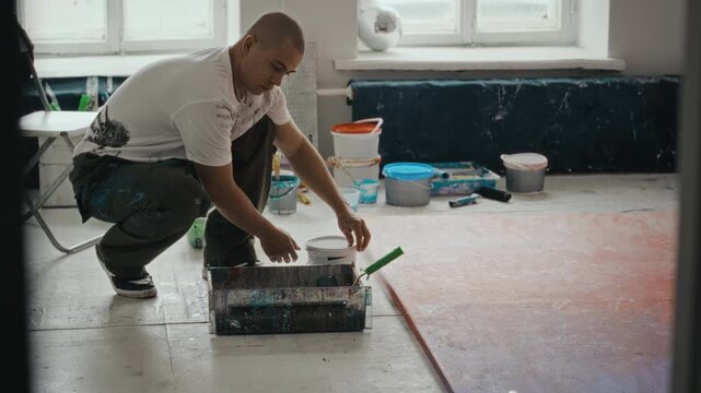 Young adult Caucasian man crouching in art studio, opening paint bucket and pouring paint into tray. Footage is ideal for creative agency promotions or art school advertisements
