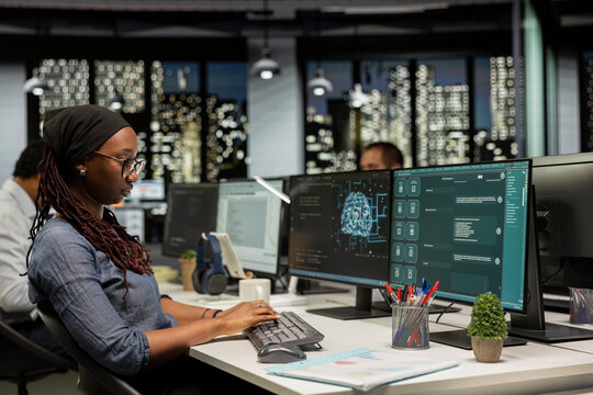 Startup worker uses a large language model application for coding help while an African American woman interacts with AI LLM chatbot software on a PC in a software development workplace.