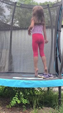 Little girl with long hair jumping on outdoor garden trampoline with safety net in backyard during summer day rear view back view concept.