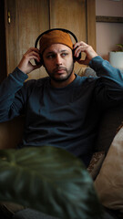 Obraz premium Young Latino man sitting on his sofa and putting on his headphones, preparing to listen to music or other audio content in a comfortable home setting.