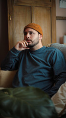 Obraz premium Young Latino man sitting on his sofa, looking very thoughtful and lost in contemplation during a quiet moment at home.
