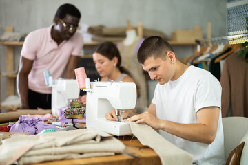 Fototapeta premium Young man working at sewing machine and friendly chatting with participants collaborating, fitting garment and preparing fabric during creative course on fashion design for adults