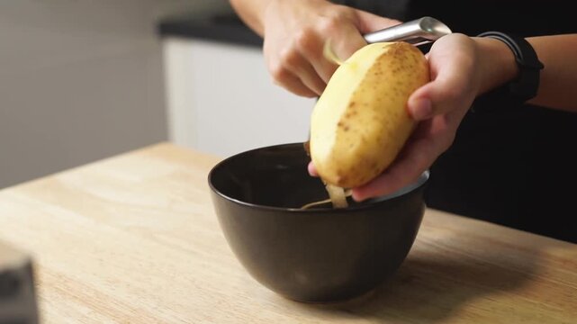 Hands peeling a potato with a peeler leaving the peels on a dark bowl. 