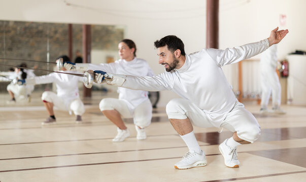 European man in white fencing uniform standing with sword in hand in front of other athletes in sports club