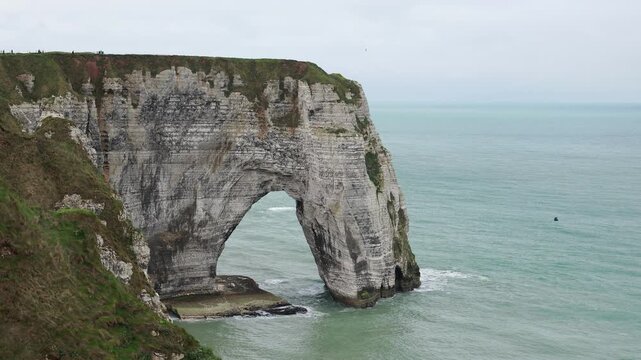 Manneporte rock arch in the chalk cliffs of Etretat, Normandy, France. Panning shot of the natural arch, layered white rock and turquoise sea on the Alabaster Coast on a grey spring day