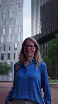 A vertical professional female wearing eyeglasses and a blue shirt looks directly at the camera outdoors. She stands in a modern city business district during a bright daytime work break