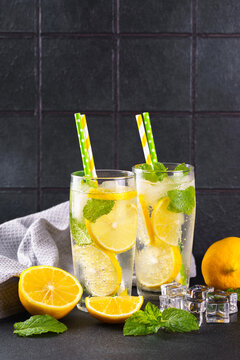 Close up of two clear glasses with refreshing homemade lemonade with fresh lemon slices, mint leaves, ice cubes and paper straws on a light kitchen counter against black tile background, copy space