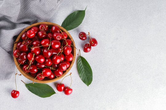 Fresh ripe sweet cherries in rustic wooden bowl with green leaves on light textured background. Top view flat lay with copy space. Juicy organic summer fruit, red berries, healthy seasonal food